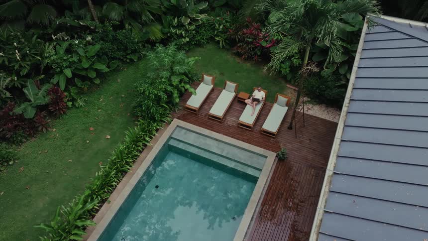 A woman relaxes on a poolside lounge chair surrounded by lush tropical greenery and a modern rooftop