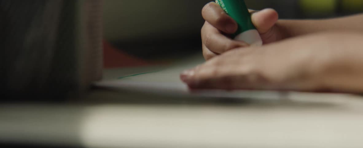 A child sits behind a desk indoors using a marker to draw on a piece of paper