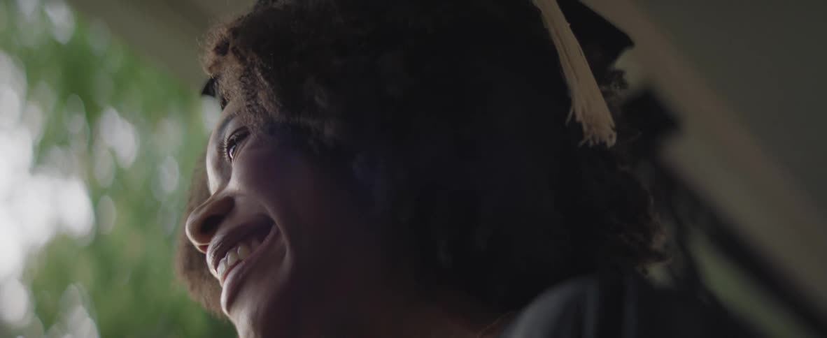 A young black woman wearing a graduation cap smiles joyfully her face lit by a soft glow from the side as she looks up and away