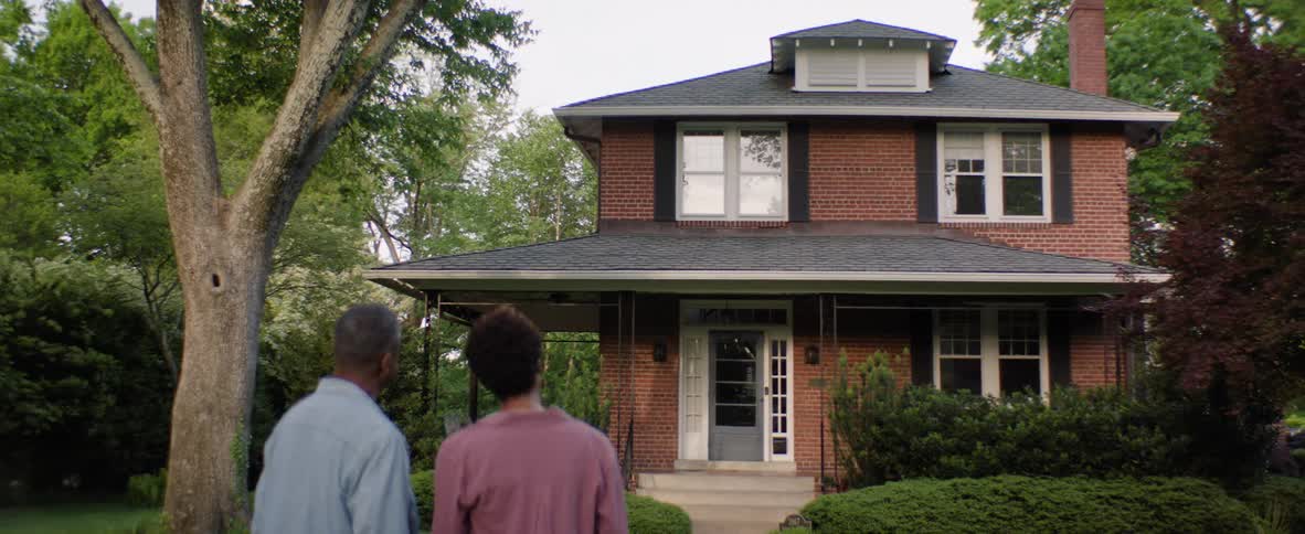 A couple stands outdoors watching a house and smiling at each other at midday