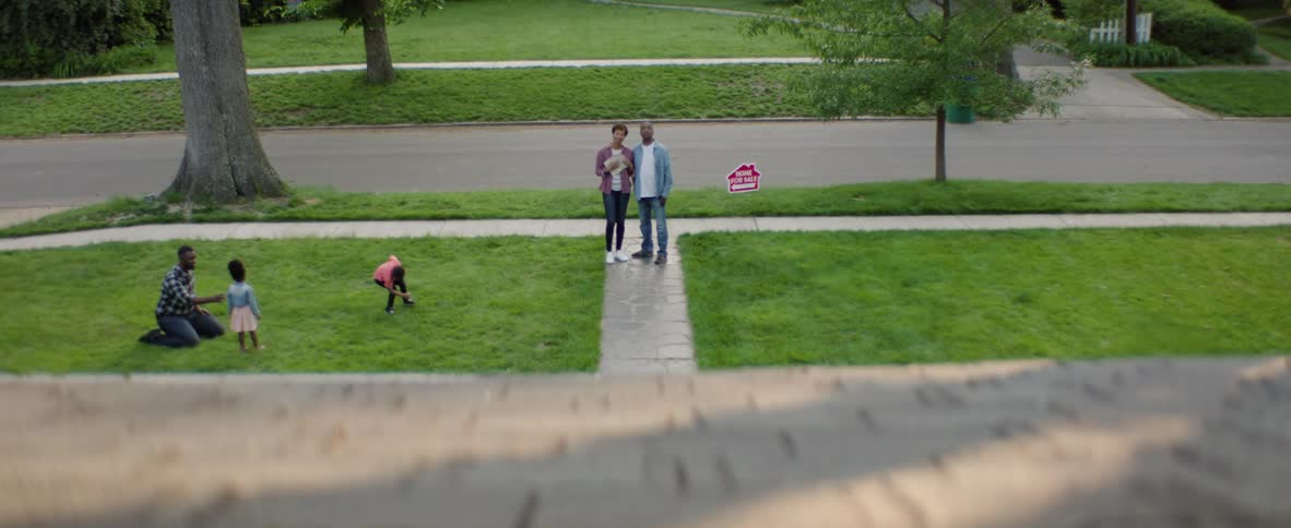 A couple stands on a paved walkway in front of a house with a home for sale sign in the yard in the foreground two young children are playing on the lawn
