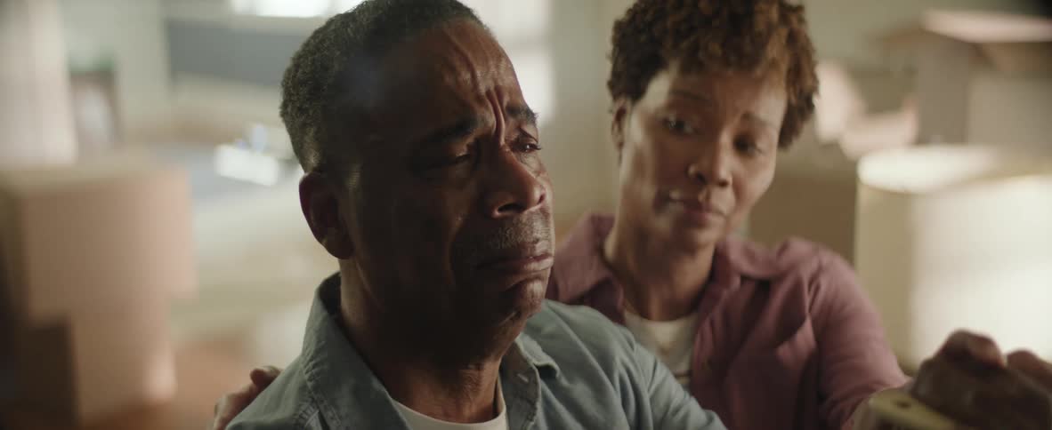A closeup shot of an emotional man is comforted by a woman who places her hand on his shoulder in a room with blurry cardboard boxes in the background