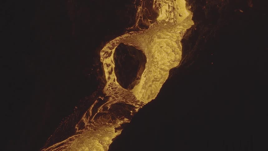 A dramatic overhead shot captures molten lava splashing violently before pulling back to reveal a glowing river of it flowing through the darkness