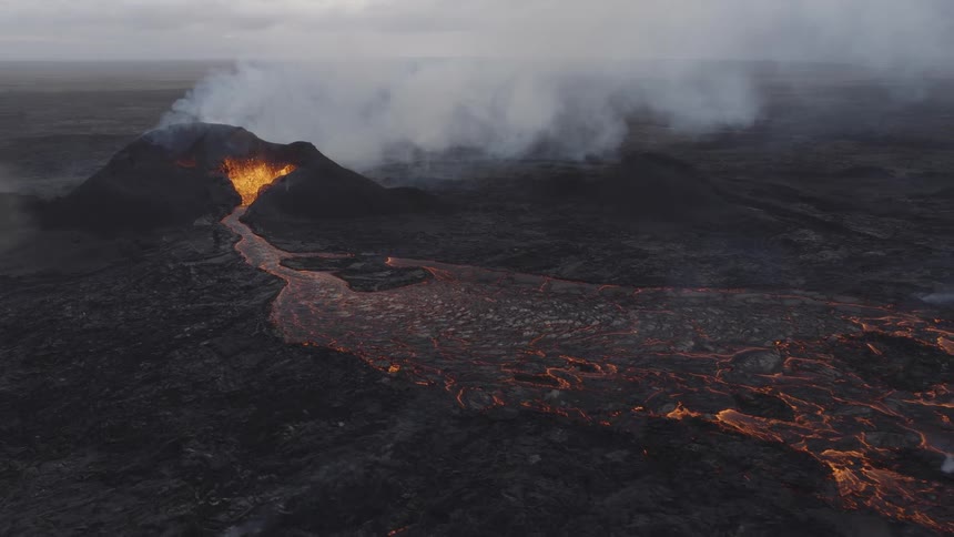 Lava flows downhill away from an erupting volcano as smoke billows up from it