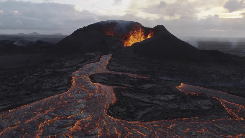 An aerial shot shows a volcano erupting as rivers of molten lava flow down its sides across a dark rocky landscape under a cloudy sky