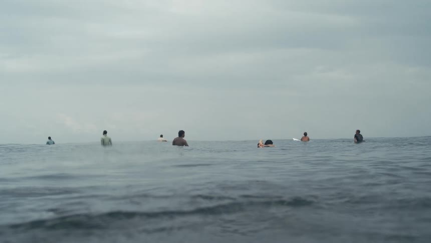 Several people float on surfboards in calm open water under a cloudy sky waiting for waves