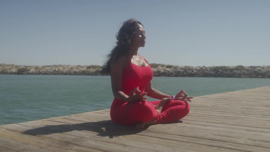 A young black woman performs yoga meditation while sitting on a wooden floor outdoors close to a river during daytime