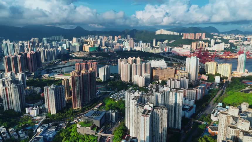 An aerial view shows a large sprawling city with numerous tall buildings and a river running through it in the distance large mountains are visible under a partly cloudy blue sky
