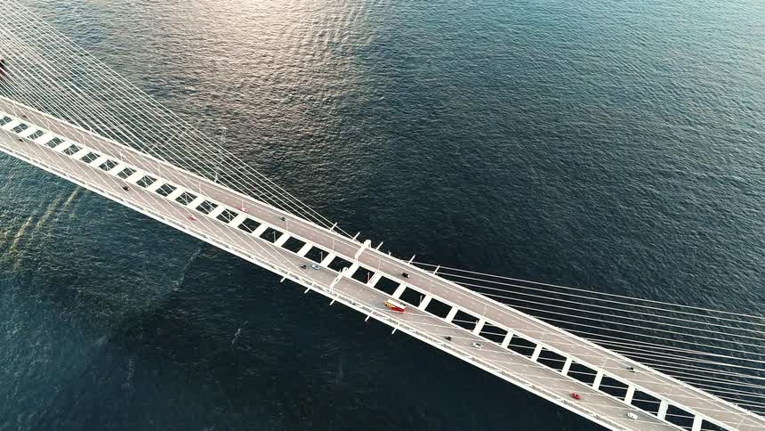 An aerial view shows traffic including cars and a bus crossing a massive white cablestayed bridge that spans a wide body of dark water
