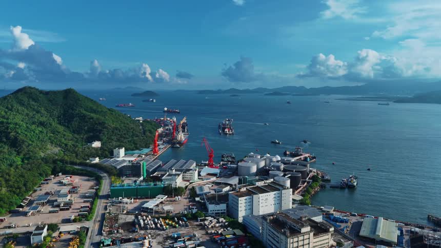 An aerial shot captures a large port or industrial area situated along a coastline with mountains in the background several boats are visible on the water and the foreground shows various buildings and a network of roads
