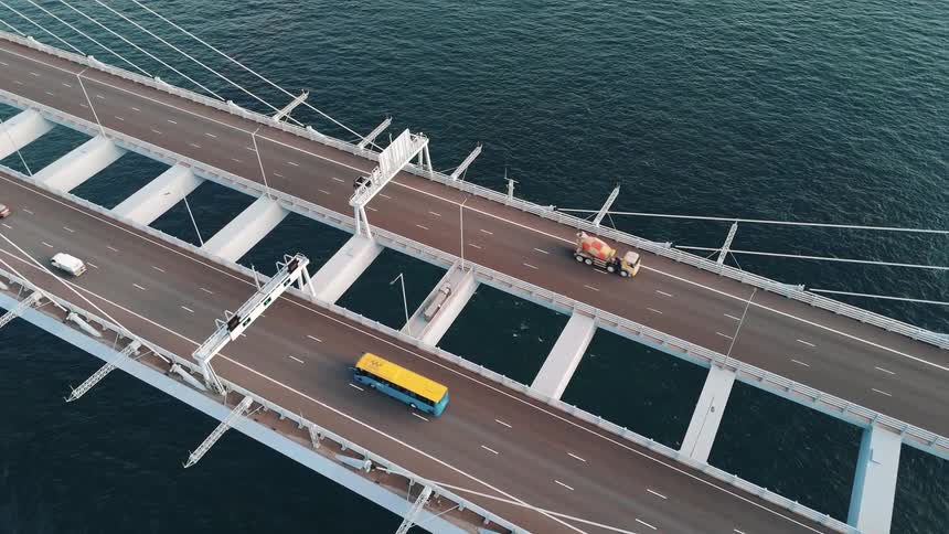 A bridge stretches across the ocean connecting two cities with cars and trucks driving by at midday