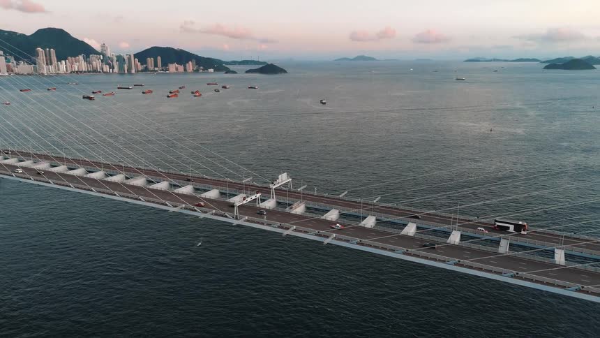An aerial shot flies alongside a massive cablestayed bridge with traffic revealing a sprawling harbor with ships and a distant city skyline at dusk