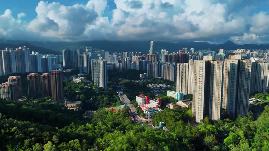 An aerial view of a city shows a series of skyscrapers surrounded by trees and green vegetation at midday