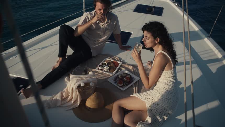 A man and woman are having a picnic on the deck of a boat with the man raising a glass to toast the woman they are sitting on blankets with a variety of snacks and the ocean is visible in the background