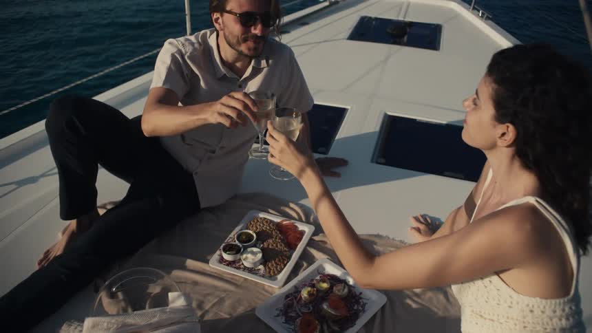 A man and woman are sitting on a boat with food and toast with a glass of wine