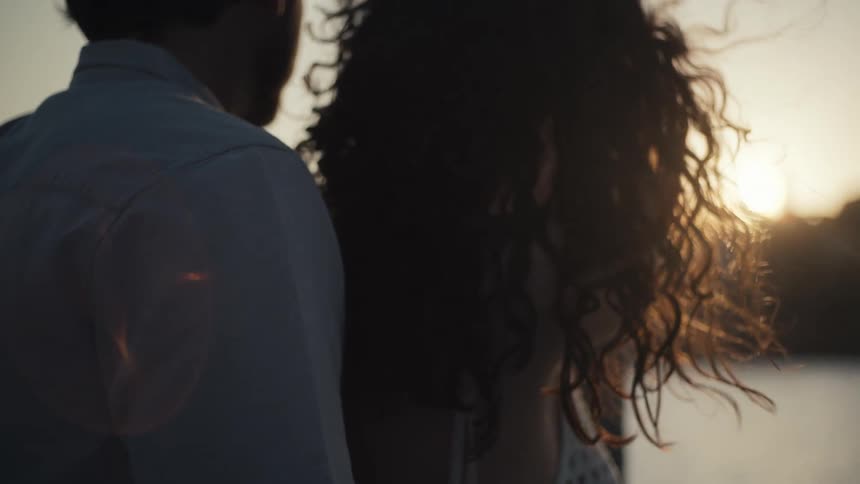 A person is standing with their back to the camera looking at the sun setting over the water they are standing close to a second person with curly hair also looking in the same direction