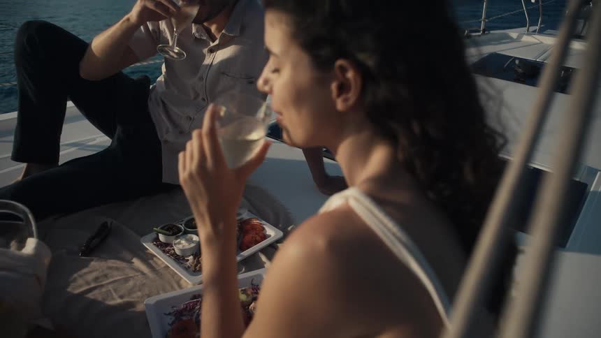 A couple is having a picnic on a boat with food and drinks the man holds up a glass to the woman who is out of focus in the foreground the couple is sitting on blankets with a variety of snacks including what appears to be olives and crackers