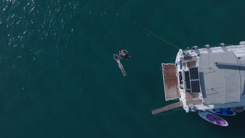 An aerial view shows two people floating in the deep blue ocean next to the back of a large white catamaran