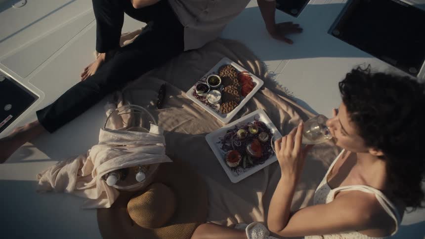 A couple enjoys a glass of wine while sitting on top of a boat during a picnic with a plate of food in front of them at midday