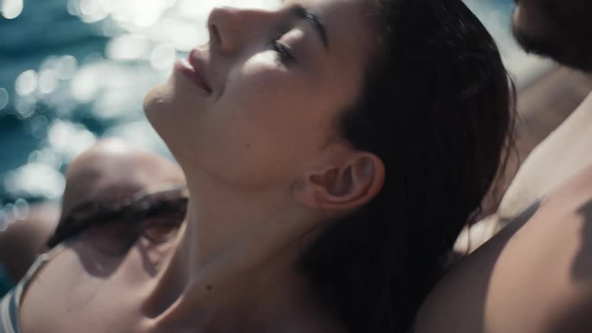 A loving couple is relaxing on a boat with the woman wearing a striped tank top leaning her head on the mans shoulder and then turning her head to look out at the sparkling blue water