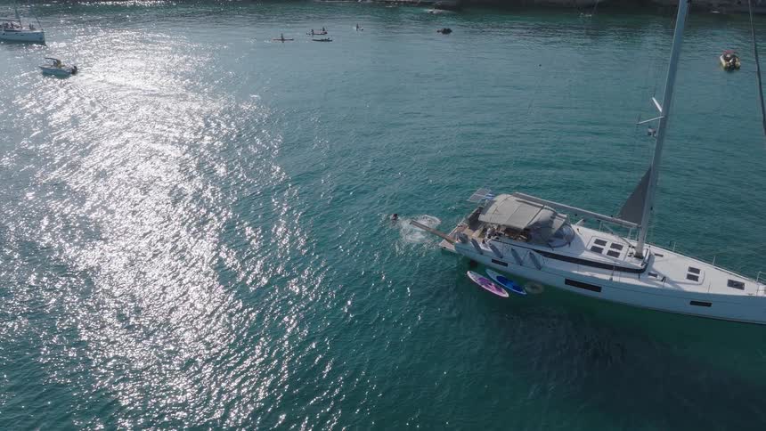 An aerial shot shows a large yacht in a clear blue bay with other boats nearby people on paddleboards are seen in the distance the water is sparkling in the sun