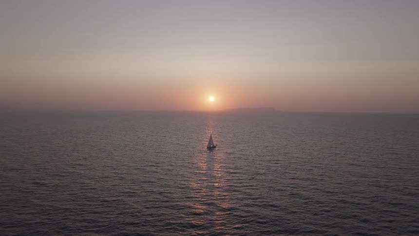 A wide aerial shot shows a single sailboat floating on a vast calm ocean at sunset