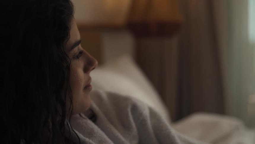 A beautiful woman with long dark hair is resting in a bed with a white blanket and a fluffy white robe looking out a window as the lamp on her bedside table illuminates her face