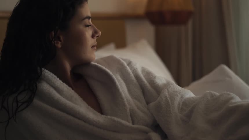 A shot shows the lower body of a person lying in bed with their hand resting on their thigh the person is wearing what appears to be a white robe and a soft filtered light is coming through a window in the background