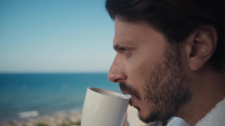 A man with a pensive expression is shown taking a sip from a mug while gazing out at a beautiful seaside view