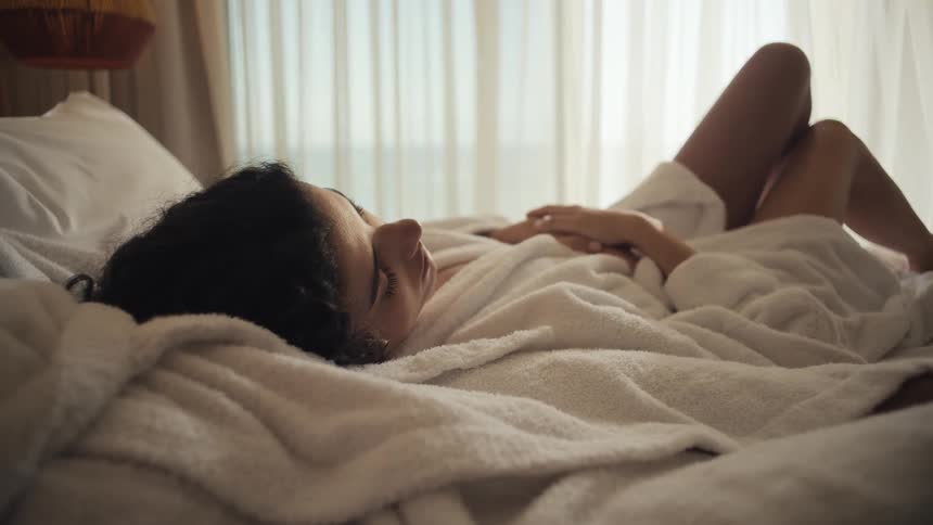 A woman with long dark hair is lying on her back in a white bed wearing a fluffy white bathrobe with her eyes closed as sunlight streams through a window behind her