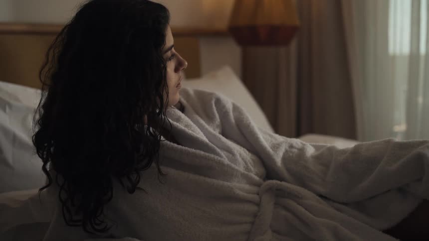 A lowangle shot shows a persons legs and midsection as they lie in bed with a white blanket covering part of their body sunlight streams in through a window in the background creating a soft hazy light