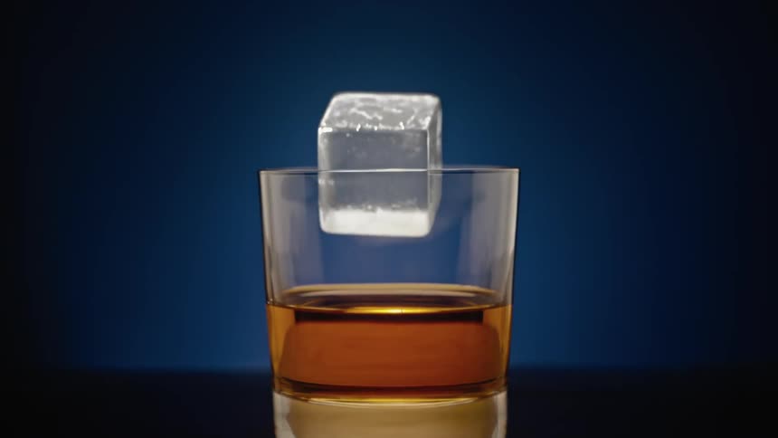 An ice cube falls and splashes into a glass of liquor