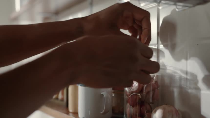 A person is putting a lid on a glass jar filled with red shallots the jar is on a wooden shelf in a kitchen next to other jars and a mug