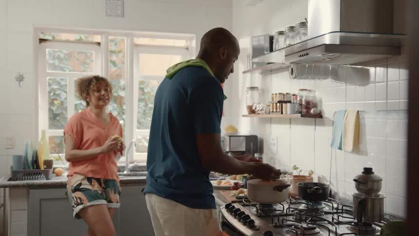 A man and a woman are in a kitchen preparing food together the man is stirring a pot on the stove while the woman stands next to him peeling an orange