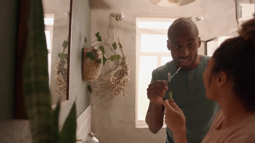 A man and a woman are in a bathroom the man is brushing his teeth while looking at the woman