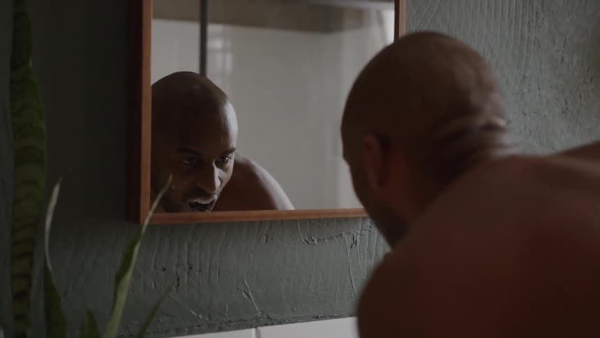 A man is looking at his reflection in a mirror while brushing his teeth he is shirtless and has a bald head