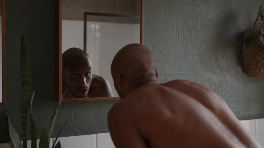A shirtless man stares intently into a bathroom mirror giving himself what appears to be a serious silent pep talk