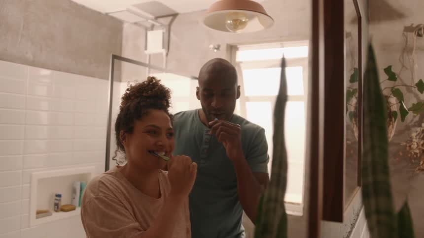 A couple is in a bathroom brushing their teeth together the woman is in the foreground and the man is standing slightly behind her
