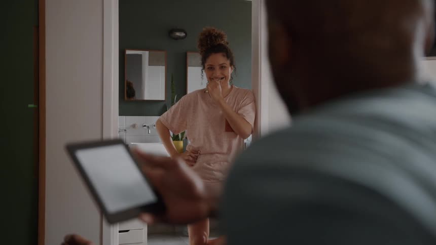 A happy woman in a tan tshirt and patterned shorts is standing in a bathroom doorway and smiling at her husband who is sitting nearby and looking at a tablet