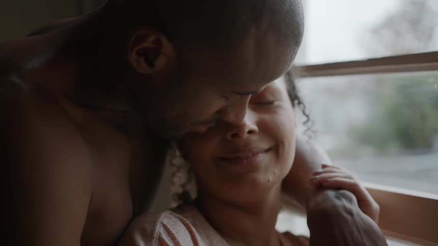 A couple is embracing near a window the man is holding the womans face and looking down at her and she is smiling