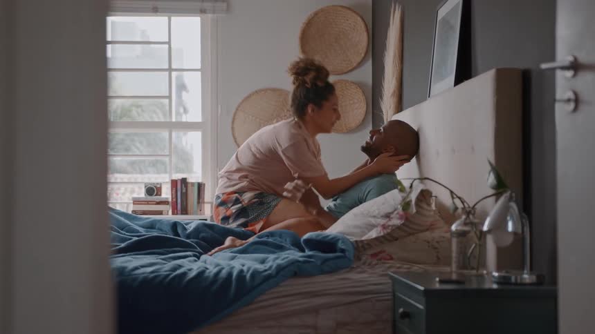 A couple is sitting on a bed in a bedroom the woman is kneeling and leaning over the man who is sitting up against the headboard