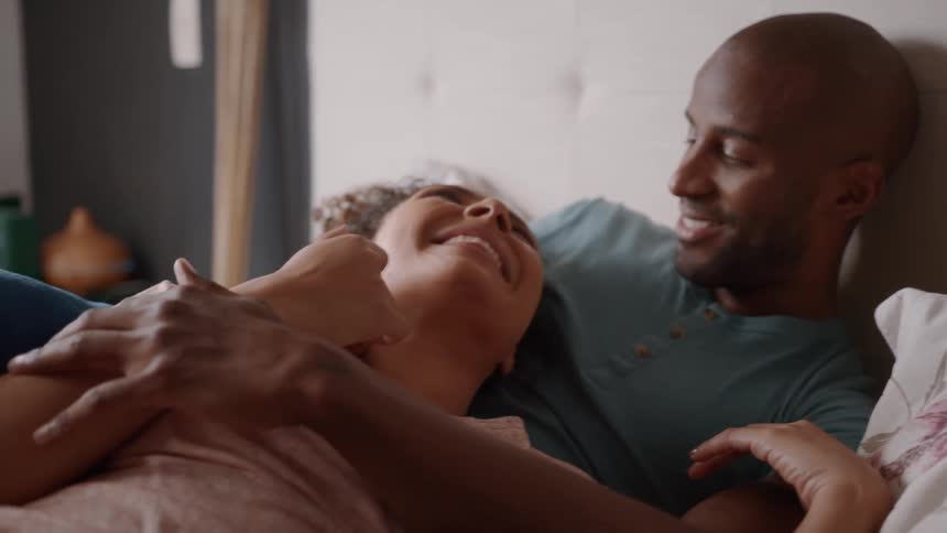 A couple lies on their bed in the bedroom with the woman resting on the mans chest as they talk and laugh enjoying a lovely time together