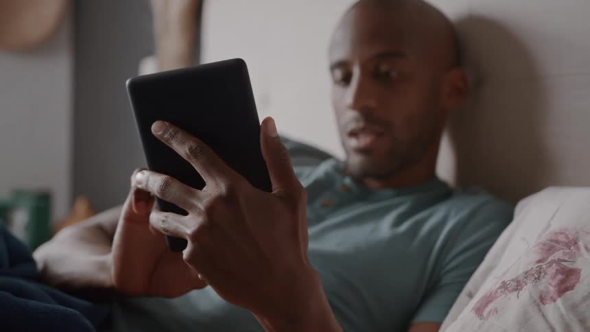 A man is shown relaxing in bed engrossed in reading something on a tablet device
