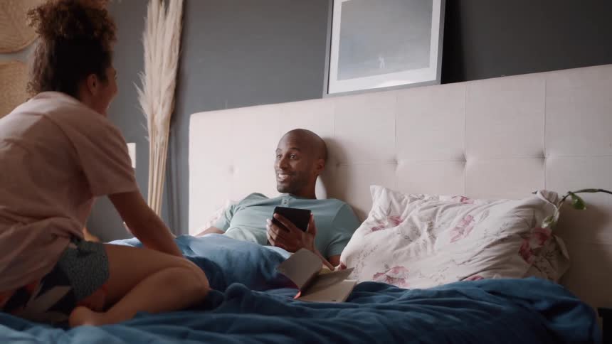 A woman reading in bed puts her book down and turns to talk to her partner who is lying beside her looking at his phone