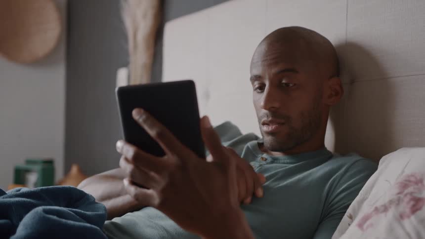 A man in bed reacting with a look of surprise and concern to something he is seeing on his tablet