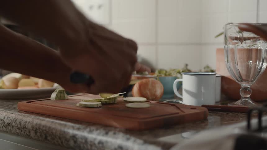 A person is transferring sliced green vegetables from a wooden cutting board in the kitchen counter with a knife into a large white pot sitting on the stovetop