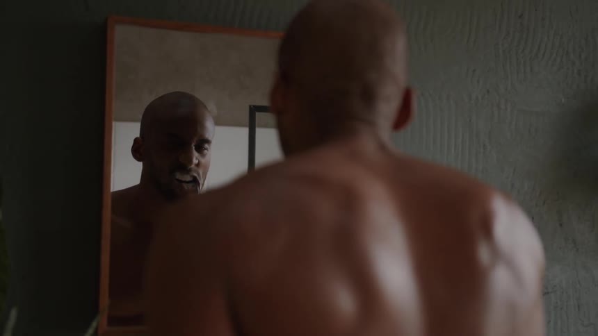 A shirtless man stands at the bathroom sink brushing his teeth