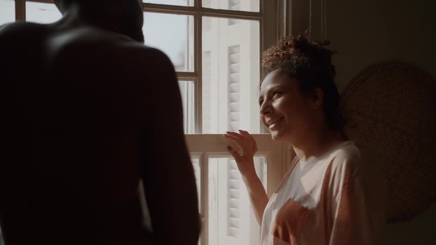 A happy couple stands next to a large window in their home with a woman in a tan tshirt talking to a man whose back is to the camera