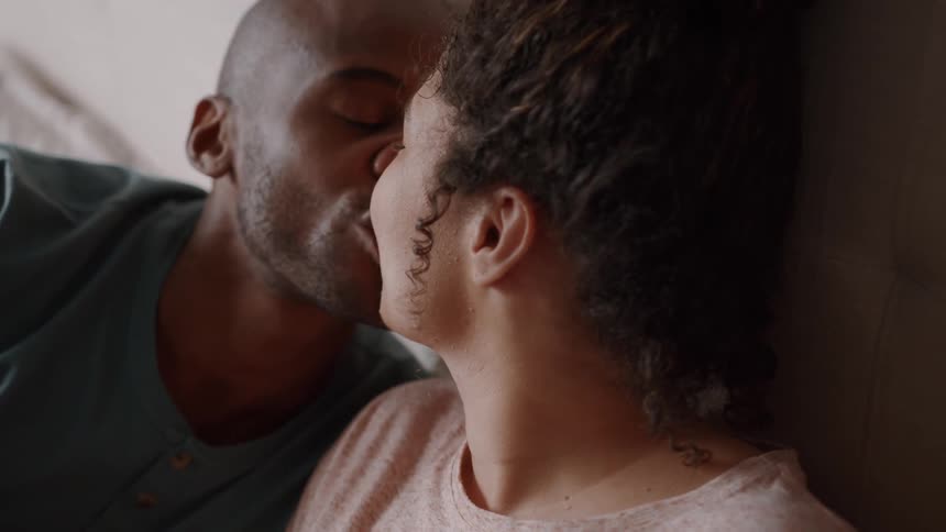 A couple sits on the floor leaning against their bed sharing a kiss in the middle of a heartfelt discussion