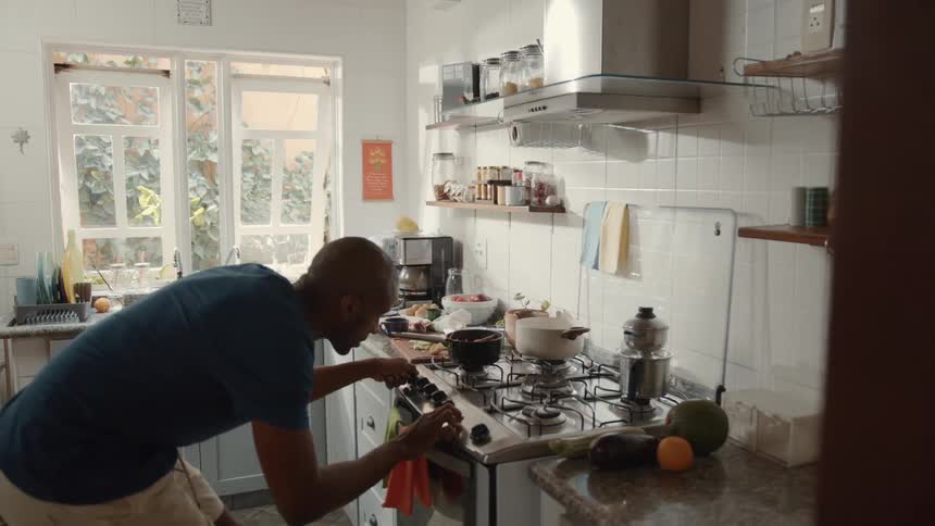 A man is in a kitchen using a pot on the stove he is focused on what hes doing and wearing a blue shirt and white pants